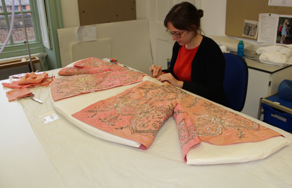 Beatrice Farmer (Senior Textile Conservator) seated at a studio table, working on the conservation of a pink watered silk jacket worn by Queen Victoria.