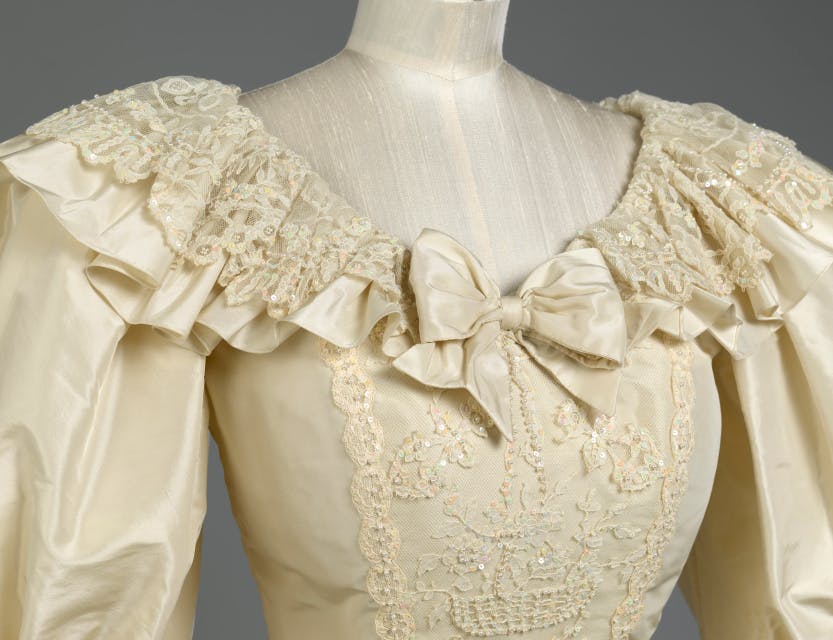 The royal wedding dress of Diana, Princess of Wales, detail. Showing the scooped neckline, part of the puffed sleeves and bodice. 

Created by designers David and Elizabeth Emanuel for Diana’s wedding to Charles, Prince of Wales on 29 July 1981. The dress is made of silk taffeta hand-embroidered with mother of pearl sequins and pearls, with a 7.5 metre detachable train. A fitted bodice is overlaid with antique carrickmacross lace, originally belonging to Queen Mary. The design features a scooped neckline, large puffed sleeves trimmed with bows and ruffles of taffeta and a full skirt. 

Royal Style in the Making explores an intimate relationship between fashion designer and royal client. Displays reveal the design process behind the creation of important one-of-a-kind couture commissions in royal history, including original sketches, fabric swatches and handwritten notes. This exhibition shows how these royal designs helped shape the British monarchy’s public image and propel the British fashion industry onto the world stage.