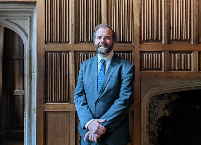 A man stands looking at the camera, in front of linenfold panelling and the corner of a Tudor door frame.