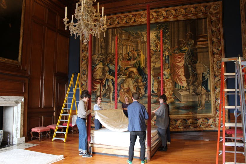 A group of people moving a large red bed frame in a room lined with tapestries