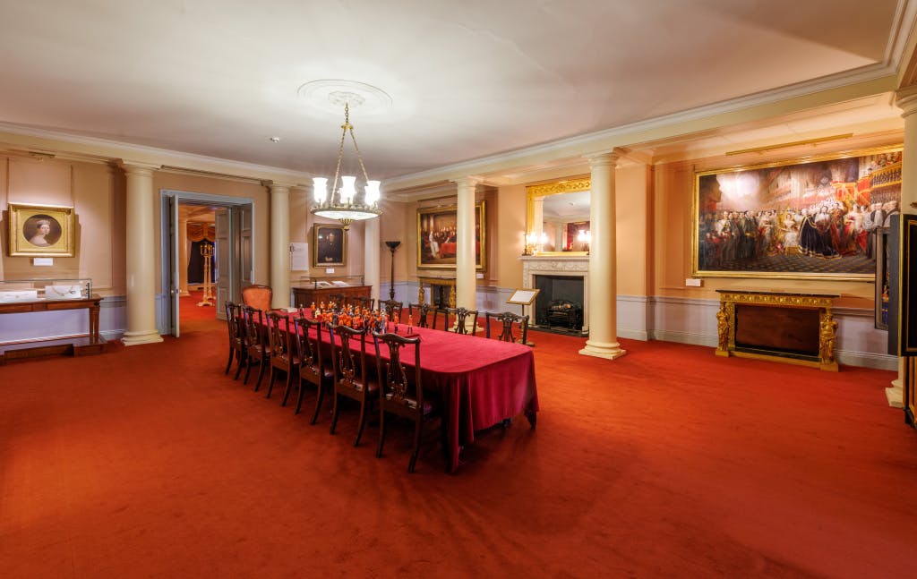 Photograph of the Red Saloon in Kensington Palace showing a long table with chairs with a striking red carpet.