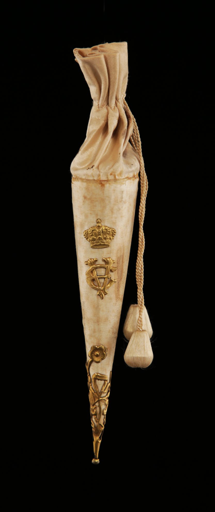 Photographed on a black background. A gold confetti bag with tassel for the wedding of Princess Victoria Eugenie of Battenberg (1887-1969).

This silk and gilt cone of rice confetti, with the initials VE adorning the side, is thought to have belonged to a bridesmaid of Victoria Eugenie of Battenberg and carried at the wedding. The confetti is still inside as tragedy struck the wedding procession when an anarchist threw a bomb at the royal carriage and an outrider was killed. Victoria Eugenia was a granddaughter of Queen Victoria. She was Queen Consort of Spain as the wife of King of Spain Alfonso XIII of Bourbon, whom she married on 31 May 1906.