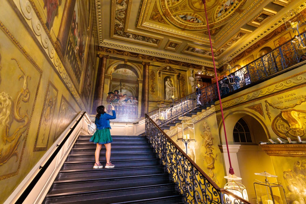 A woman takes a photo of a grand, golden staircase with a decorative mural on the wall