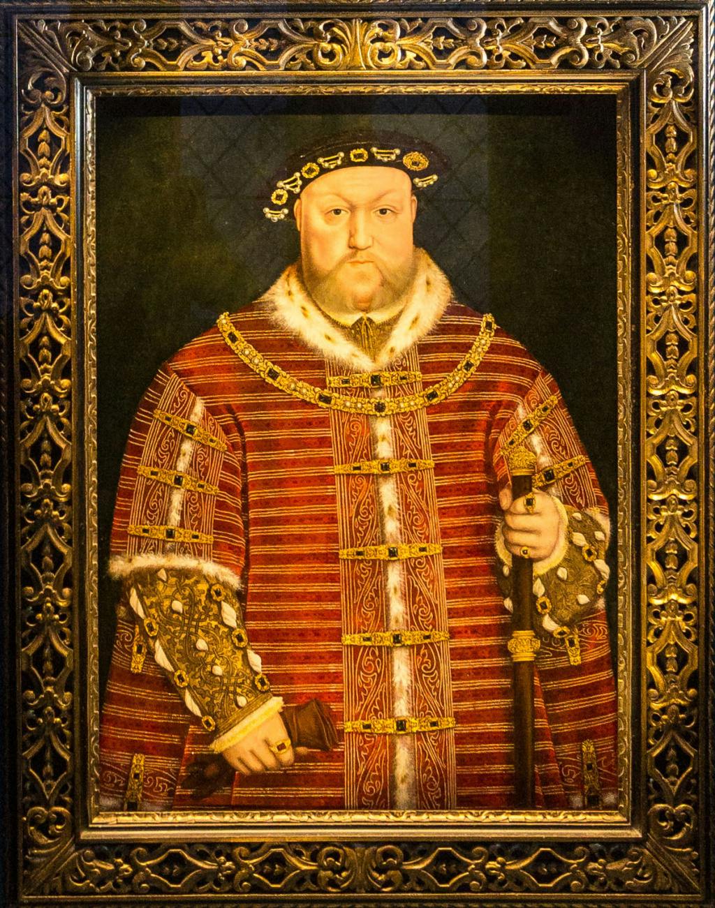 Henry VIII portrait at Hampton Court Palace, showing the King facing the viewer, wearing a deep red gown, lined with fur and decorated with jewels. He holds in his left hand a wooden staff in his hand adorned with gold, and in his left hand he holds a glove.
