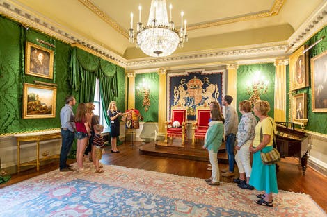 Гид показывает группе посетителей Тронный зал в парадных залах замка и садов Хиллсборо. Два красно-золотых трона стоят в одном конце комнаты, а стены обшиты зеленым шелковым дамаском, а стены украшены историческими произведениями искусства.