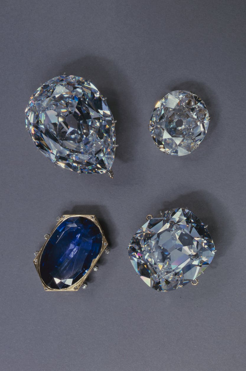 A portrait photograph of The Cullinan I and Cullinan II diamonds, the Koh-i-Noor diamond and the Stuart Sapphire from the Crown Jewels. displayed against a grey background.