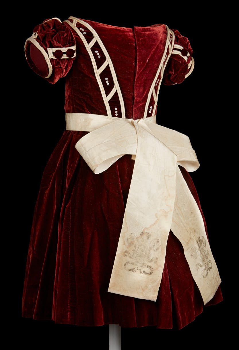 A rear view of a burgundy dress photographed against a black background. The dress is made from burgundy velvet with ivory satin piping, mother of pearl buttons, and a taffeta waist sash