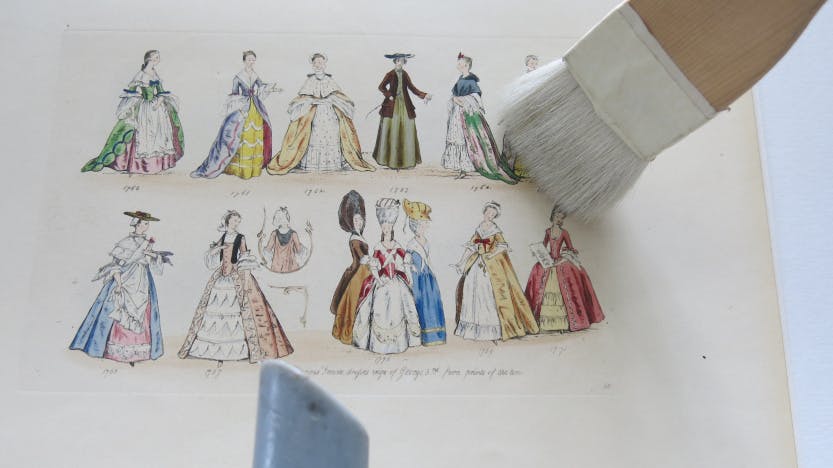 Close up of a brush being used to remove dust from a fashion plate. "Female Dress from 1760-1776", engraved by Charles and Leopold Martin and published in 1840. Shows two rows of ladies in different fashion dress.