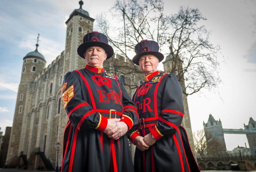 Two Yeoman Warders, also known as Beefeaters, stand in front of the White Tower of the Tower of London on a cloudy day