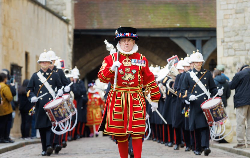 Chief Yeoman Warder Rob Fuller parades in front of a Royal Marines military band, flanked by drummers on either side of him, He wears traditional Yeoman Warder State Dress. His red tunic has gold accents and the initials of the monarch on his chest, and he carries a silver mace topped with a model of the White Tower over his shoulder.