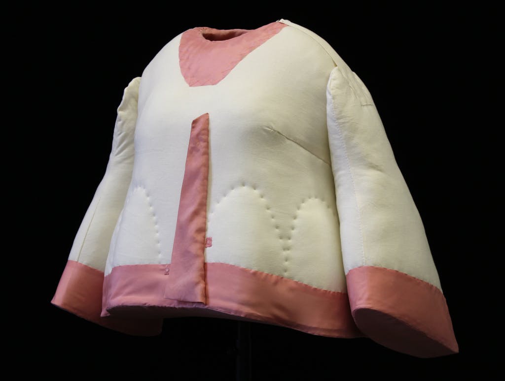 A cutaway acrylic mannequin created by Hampton Court textile conservators to display a jacket worn by Queen Victoria. The mannequin is lined in pink silk (intended to look like the lining of the jacket) and the smooth acrylic covered with calico to allow padding to be stitched on. Mannequin photographed against a black background.