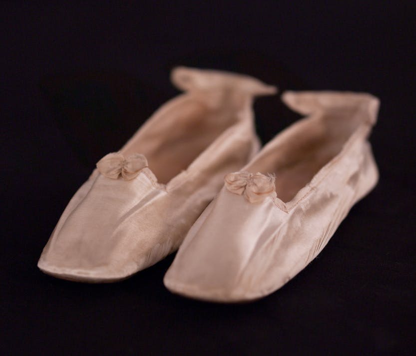 A pair of ivory silk satin shoes believed to have been worn by Princess Beatrice (1857-1944) when she was about 18 months old. Photographed on a black background.