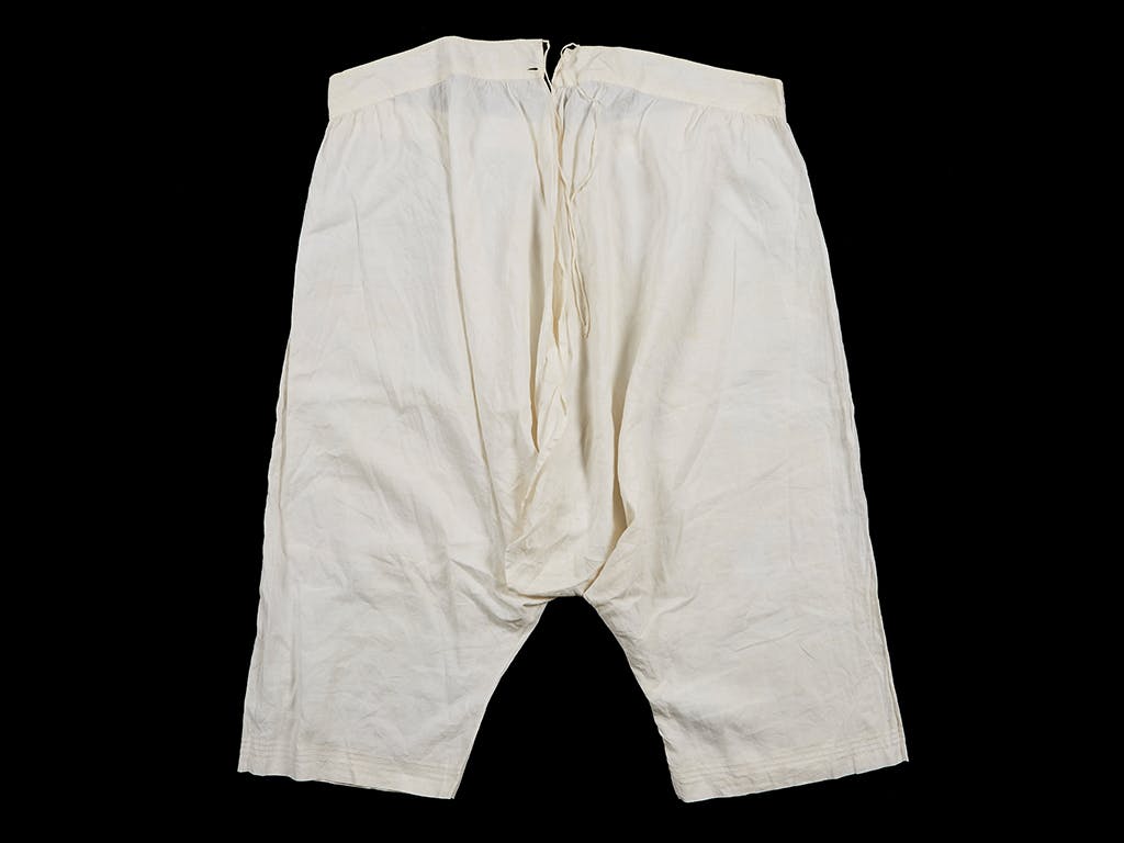 A pair of just below knee-length, white cotton open drawers or knickers worn by Queen Victoria (1819-1901). Photographed against a black background.