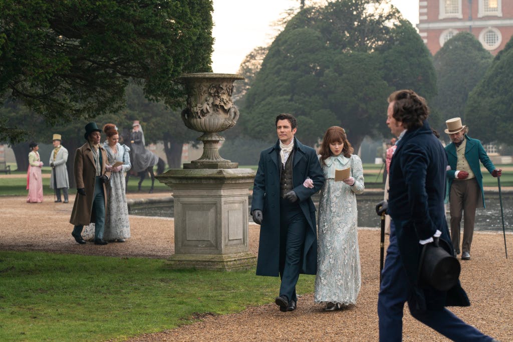 A group of people dressed in regency costume walking through the gardens at Hampton Court Palace.