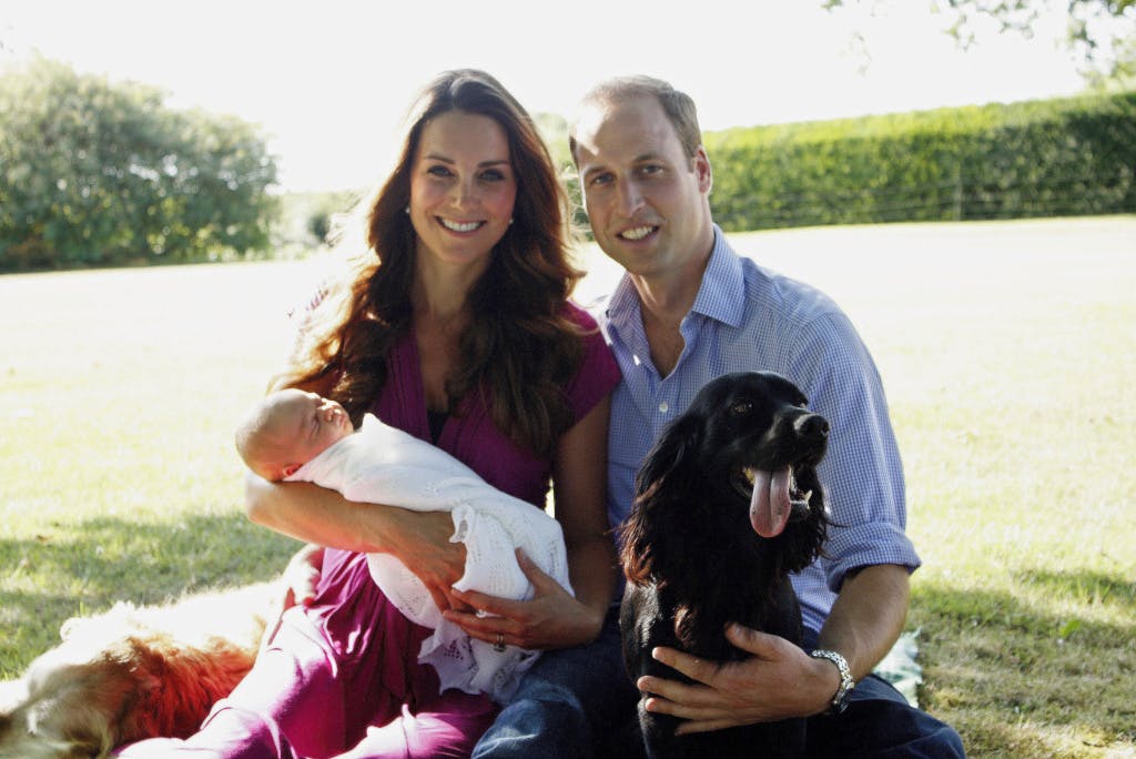 TRH The Prince and Princess of Wales sit on the ground in a green garden, smiling at the camera. The Princess holds Prince George as a baby in her arms. A black dog sits next to the Prince of Wales.