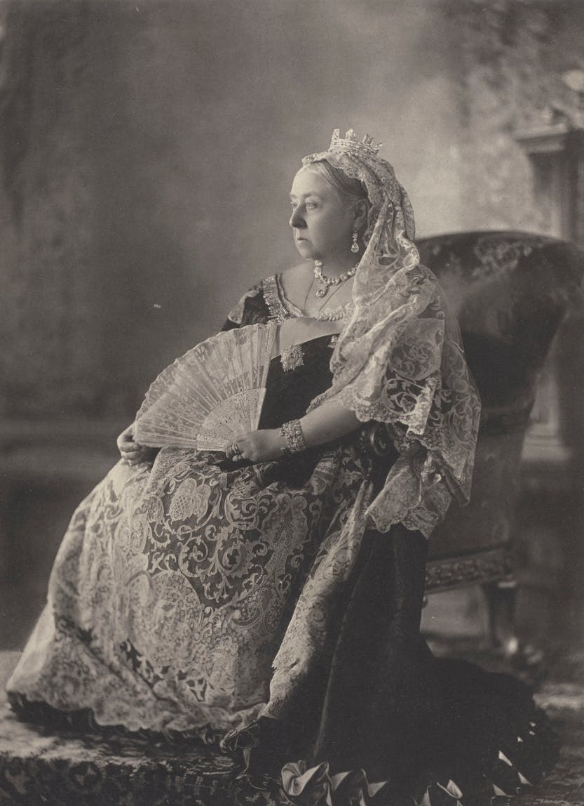 Photograph of Queen Victoria, full length, seated and facing three-quarters right, dressed in lace. She holds a fan in her left hand.