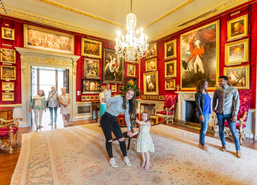 A group of visitors enjoying a viewing of the Red Room, one of Hillsborough Castle's State Rooms. Two young girls appear in the foreground along with a young couple. A small group of older ladies and gentlemen can be seen in the background having just entered the room. The walls are lined with a deep red silk damask and a variety of oil paintings in gold frames of various sizes fill the walls. A chandelier hangs above a small round wooden table in the centre of the room. The walls are lined with grey and gold detailing.