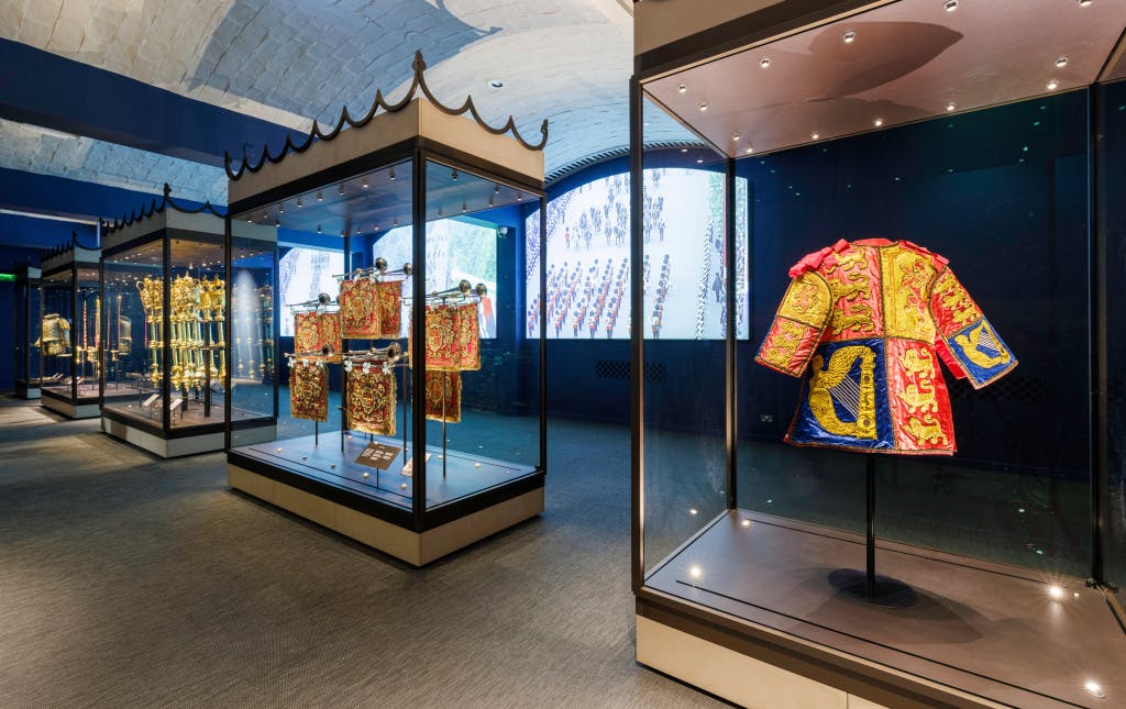 The New Jewel House exhibition, showing a general view of Room 4 - Procession (c). Objects displayed include the Richmond Herald tabard, part of the Court Dress uniforms of Sir Gerald Woods Woolaston KCB, KCVO (1874-1957). The room is spacious with large display cases.