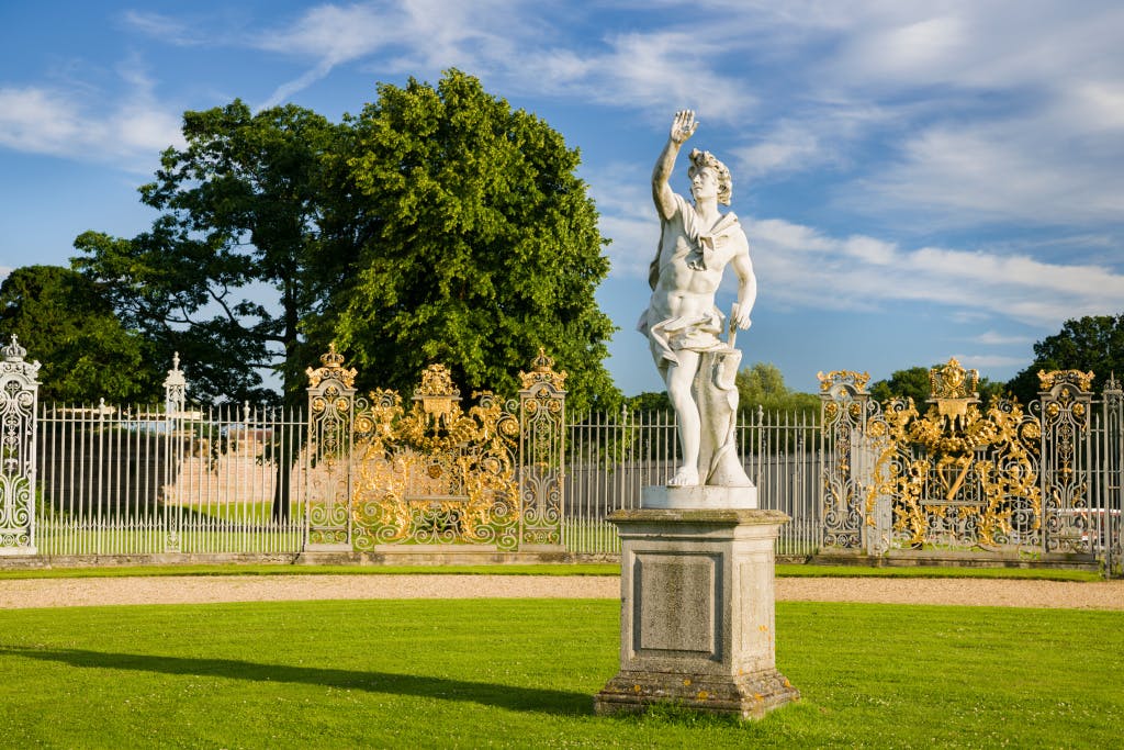A formal garden with a white statue in the foreground and a ironwork gold screen behind