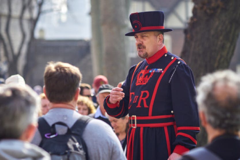 A Yeoman Warder in full uniform giving a tour of the Tower of London to a group of visitors. Tudor and medieval buildings can be seen in the background