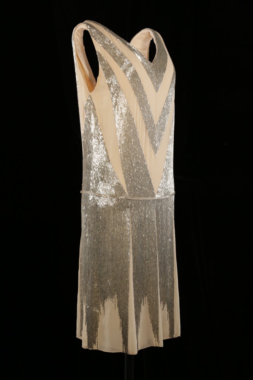 Front side view of a sleeveless, drop-waist court dress of pale peach silk georgette with silver bugle beads. Created by Jeanne Lanvin (1867-1946).

This dress was worn by Lillian Wardman at her daughter’s court presentation ceremony in 1928.

Photographed on a black background.