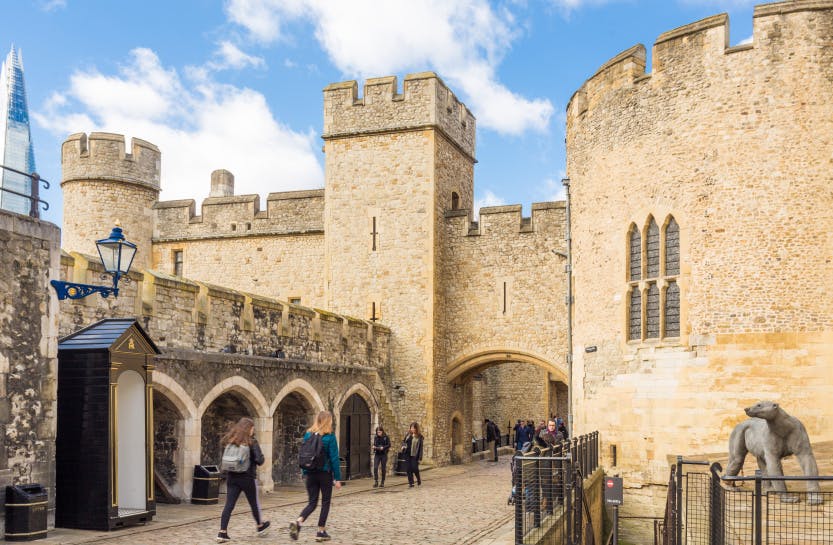 The stone Wakefield Tower and visitor exit at the Tower of London under a blue partially cloudy sky, showing visitors exploring the grounds below the tower