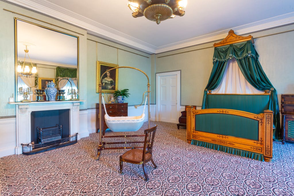 Victorian period furniture in a green room. There is a patterned carpet on the floor and a large bed, belonging to Queen Victoria and Prince Albert, in the far end of the room