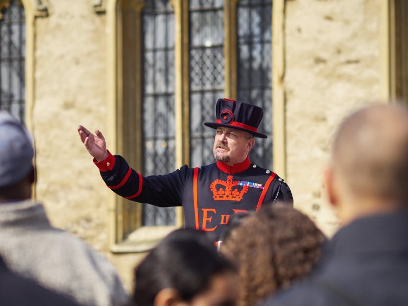 A Yeoman Warder is shown in their full uniform speaking to a group of visitors.  One arm is raised.